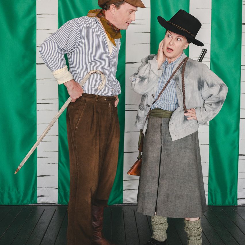 Two individuals dressed in vintage attire, one holding a cane and the other with a rifle, stand on a stage with a green and white backdrop.