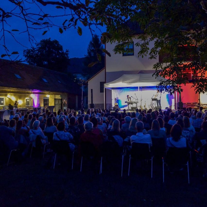 A nighttime outdoor concert with a stage under a tent, featuring two performers playing instruments. The audience is seated in chairs, illuminated by colored lights.