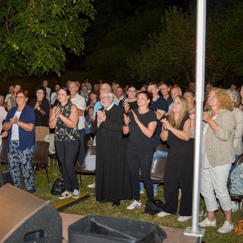 A large group of people, including a priest, are gathered in an outdoor area at night, clapping and smiling, with trees and a stage in the background.