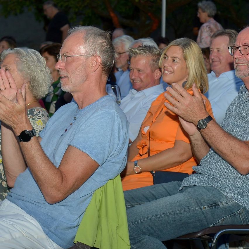 A group of people are sitting in a row, clapping their hands. The man in the blue shirt is wearing glasses and a watch.