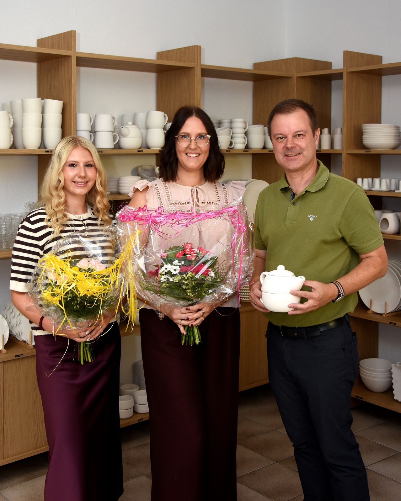 Drei Personen stehen in einem Raum mit Regalen voller weißer Geschirr, zwei Frauen halten Blumensträuße und ein Mann hält eine Teekanne.