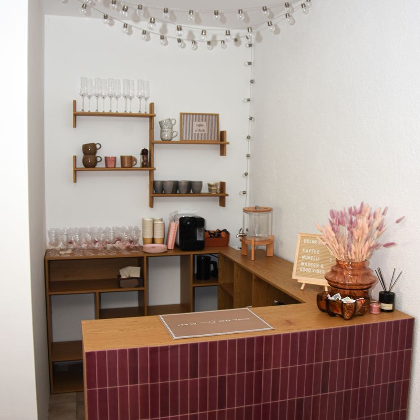 Ein kleiner, gemütlicher Café mit Holzregalen, einer Theke und dekorativen Gegenständen. Die Theke hat ein Schild mit der Aufschrift 'Getränke, Kaffee, Murelli, Wasser & Gute Laune.' Auf den Regalen stehen Weingläser, Tassen und eine Kaffeemaschine.
