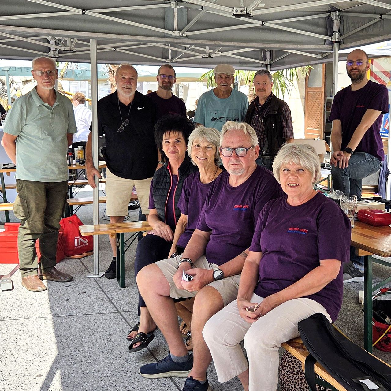 Eine Gruppe von Menschen in lila T-Shirts sitzt auf einer Bank unter einem Zelt. Sie lächeln und posieren für ein Foto. Dahinter steht ein Tisch mit einem Glas und einer roten Tasche. Links steht ein Mann und schaut sich etwas an.