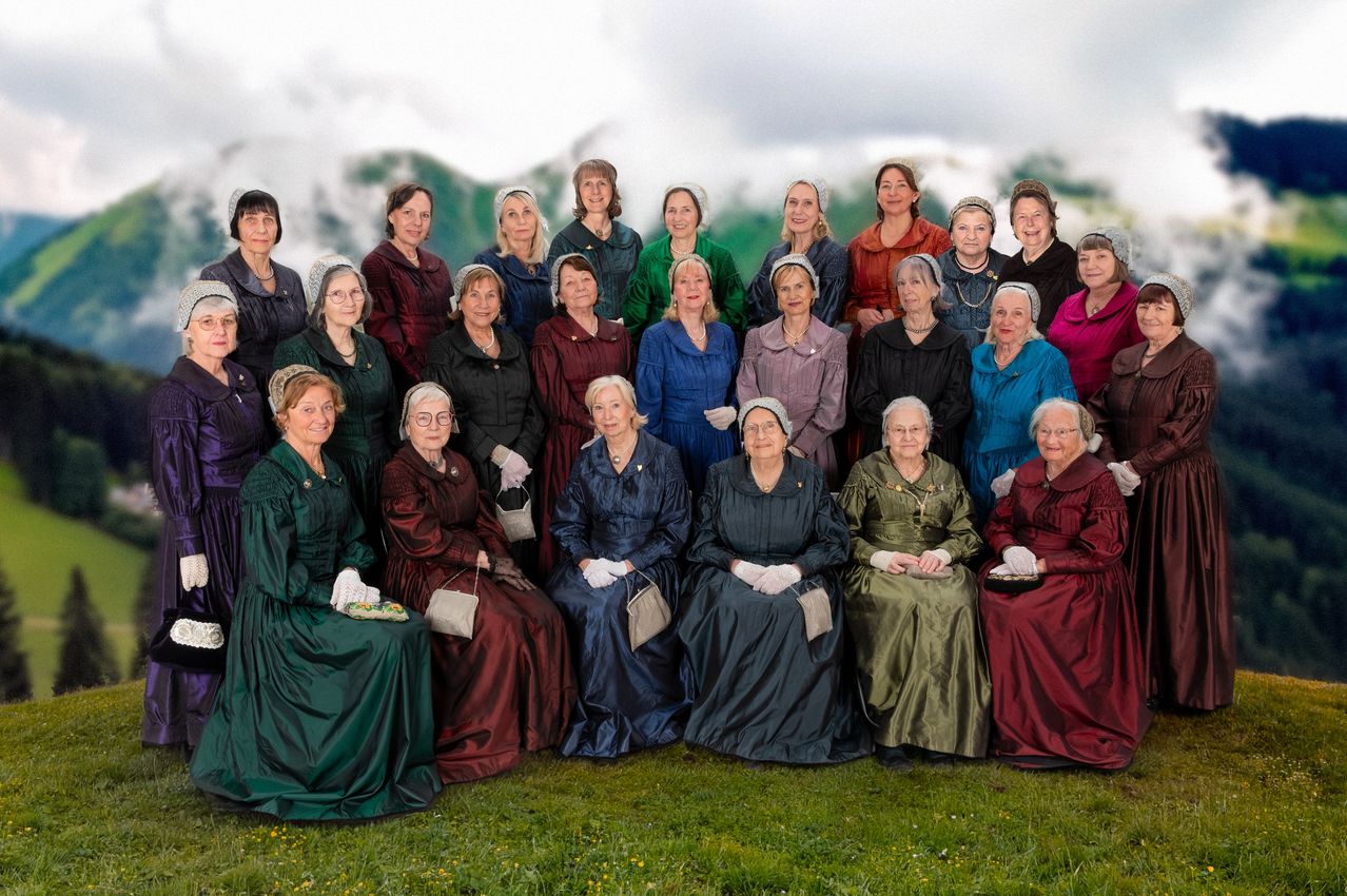Eine Gruppe von Frauen in viktorianischer Kleidung, die vor einem Berghintergrund sitzen und stehen. Sie tragen Hauben und Handschuhe und einige halten kleine Taschen.