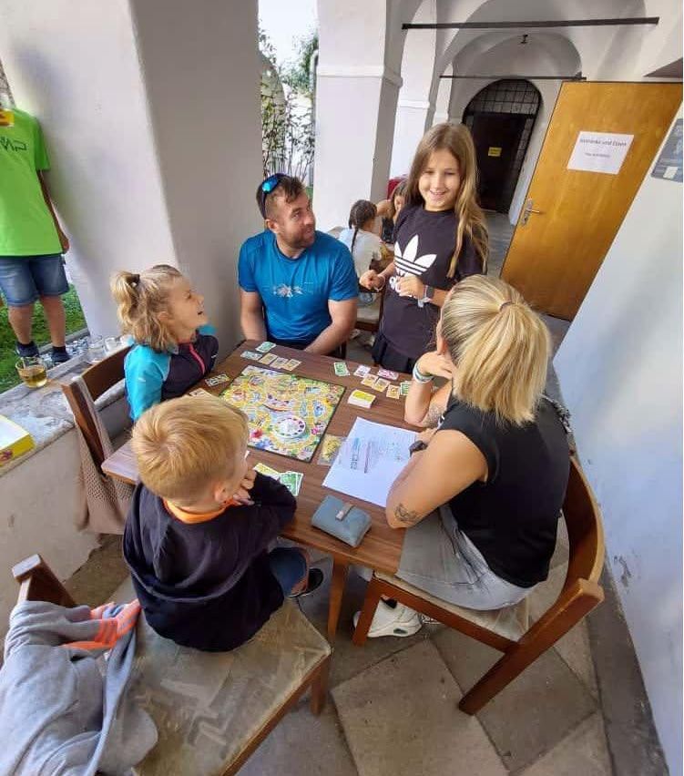 Eine Familie sitzt um einen Tisch herum und spielt ein Brettspiel, wobei eine Frau und zwei Mädchen in der Nähe stehen.