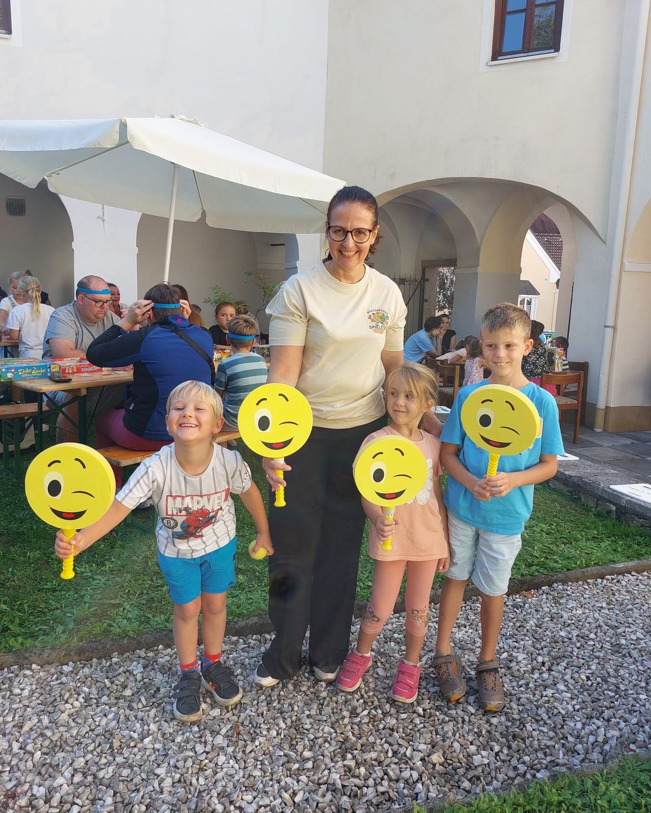 Eine Frau und drei Kinder stehen zusammen und halten gelbe Smiley-Gesichter-Schilder, lächeln für ein Foto in einem Garten. Hinter ihnen sitzen Menschen an Tischen, und ein Schirm ist in der Nähe.