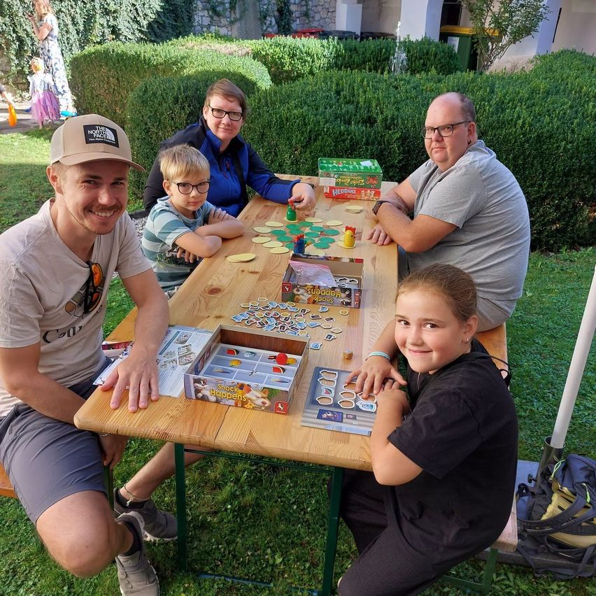 Eine Familie sitzt an einem Tisch und spielt an einem sonnigen Tag ein Brettspiel.