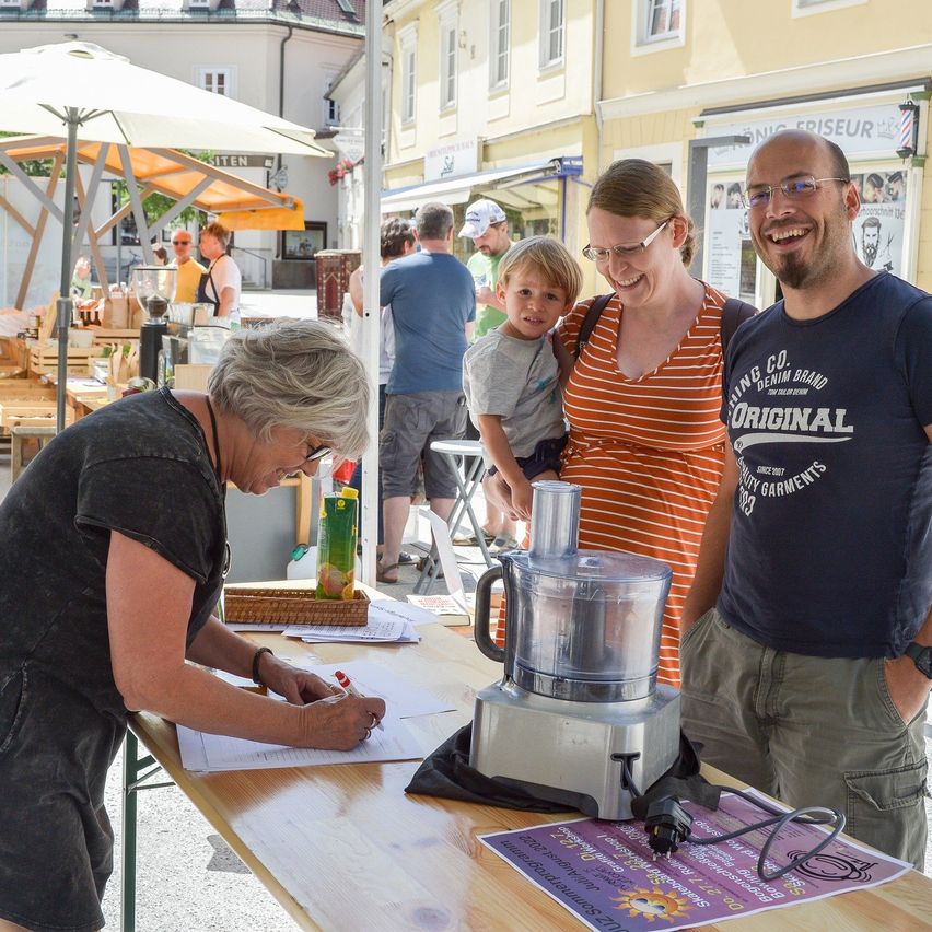Eine Frau unterschreibt ein Dokument an einem Tisch mit einem Mixer und einer Orangensaftbox. In der Nähe hält ein lächelndes Paar ein Kind. Dahinter ein Markt mit Schirmen und Gebäuden.