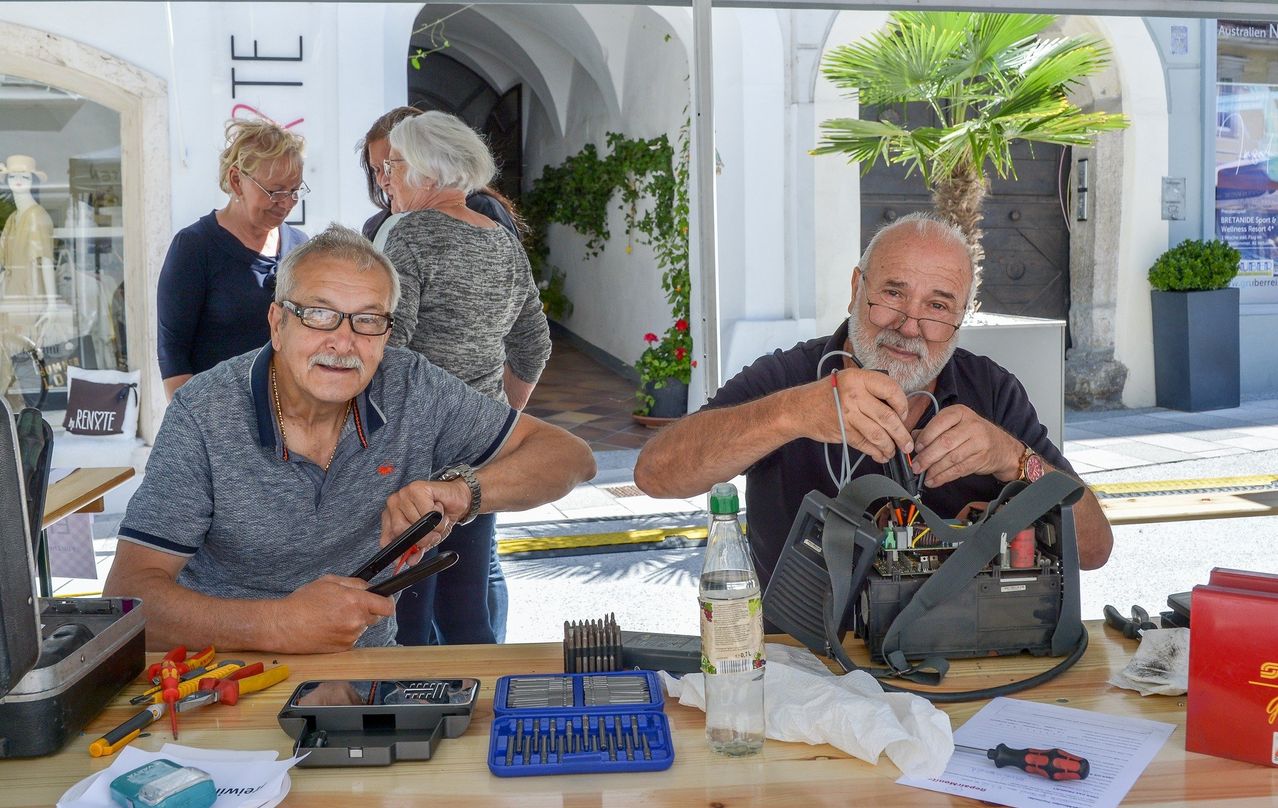 Zwei ältere Männer mit grauen Haaren und Brille arbeiten an einem elektronischen Gerät auf einem Tisch. Verschiedene Werkzeuge und eine Flasche befinden sich auf dem Tisch. Hinter ihnen beobachtet eine Frau ihre Arbeit.