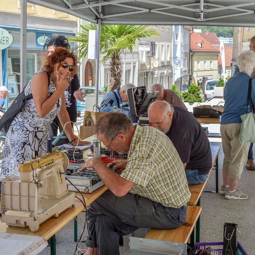 Ein Freiluftmarkt, wo ein älterer Mann an einer Maschine arbeitet, während eine Frau mit Sonnenbrille steht und mehrere Leute sich um einen anderen Mann versammelt haben, der einen geöffneten Laptop repariert.