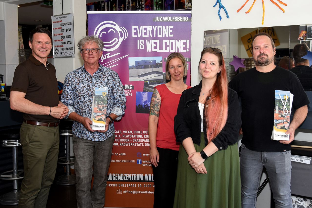 Fünf Personen stehen vor einem Banner, einer Mann hält ein Buch. Dahinter steht ein Banner mit der Aufschrift 'Jeder ist willkommen!' und einem Logo. Zwei Frauen und zwei Männer lächeln in die Kamera.