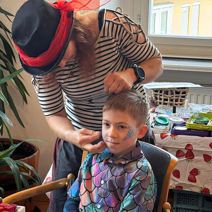 Eine Frau malt ein Design auf das Gesicht eines Jungen, der auf einem Stuhl sitzt. Eine Pflanze ist links.