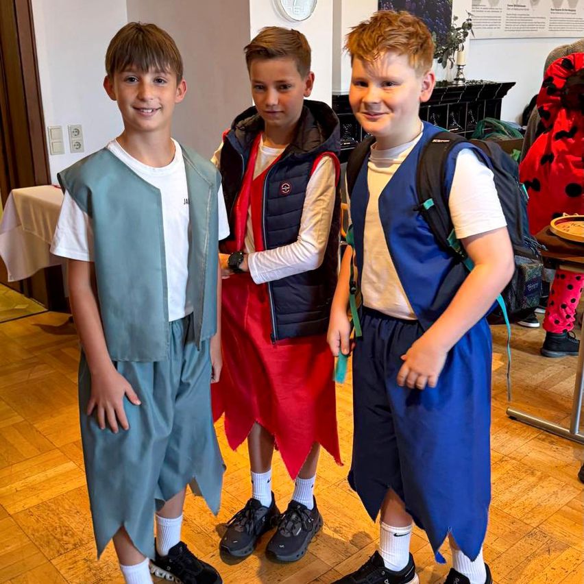Drei Jungen in Kostümen stehen lächelnd für die Kamera auf einem Holzboden. Der Junge links trägt eine blaue Weste, der in der Mitte ein rotes Kleid und der Junge rechts eine blaue Weste mit Rucksack.