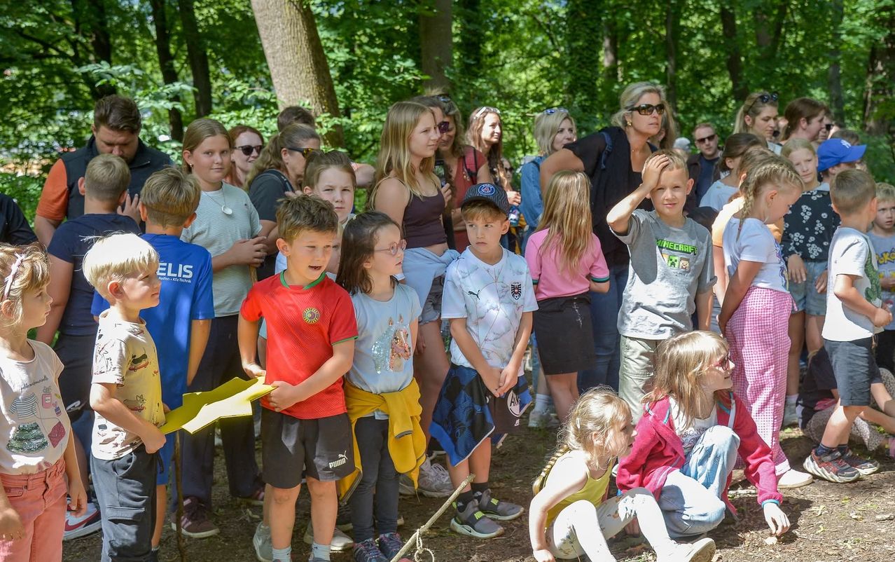 Eine Gruppe von Kindern und Erwachsenen steht und sitzt in einem bewaldeten Gebiet, wahrscheinlich an einem sonnigen Tag. Einige tragen Brillen und Turnschuhe, während ein Kind ein gelbes Papier hält.