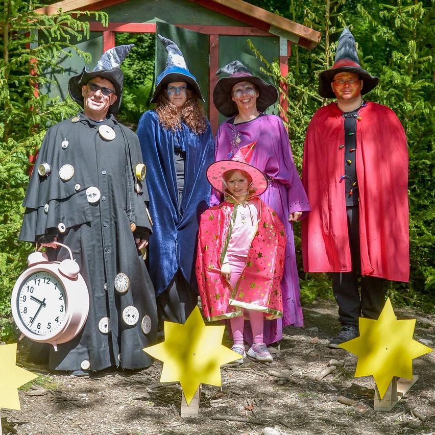 Fünf Personen in Hexenkostümen stehen in einem Wald mit gelben Sternen auf dem Boden. Einer hält eine Uhr. Dahinter steht ein kleines Haus mit einer Tür.