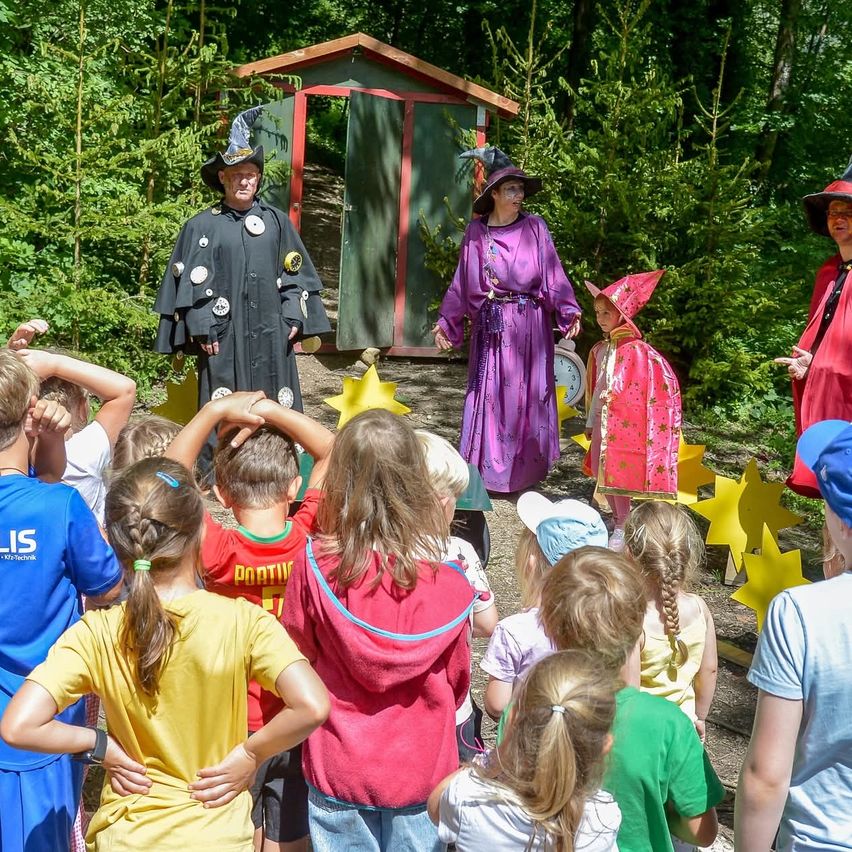 Mehrere Erwachsene und Kinder sind im Freien, wobei Erwachsene in Kostümen ein Theaterstück aufführen. Das Publikum der Kinder schaut aufmerksam zu.