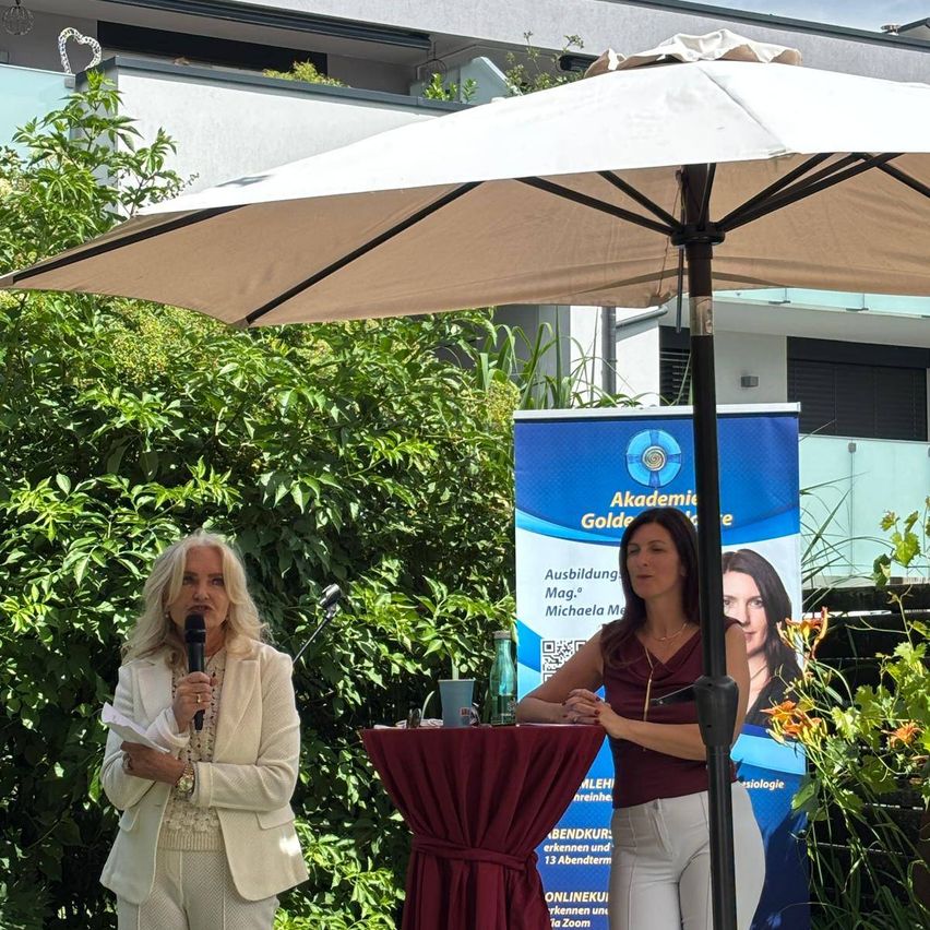 Zwei Frauen sprechen in Mikrofone unter einem Regenschirm. Dahinter befindet sich ein Banner für Akademie Golden, das einen Kurs bewirbt.