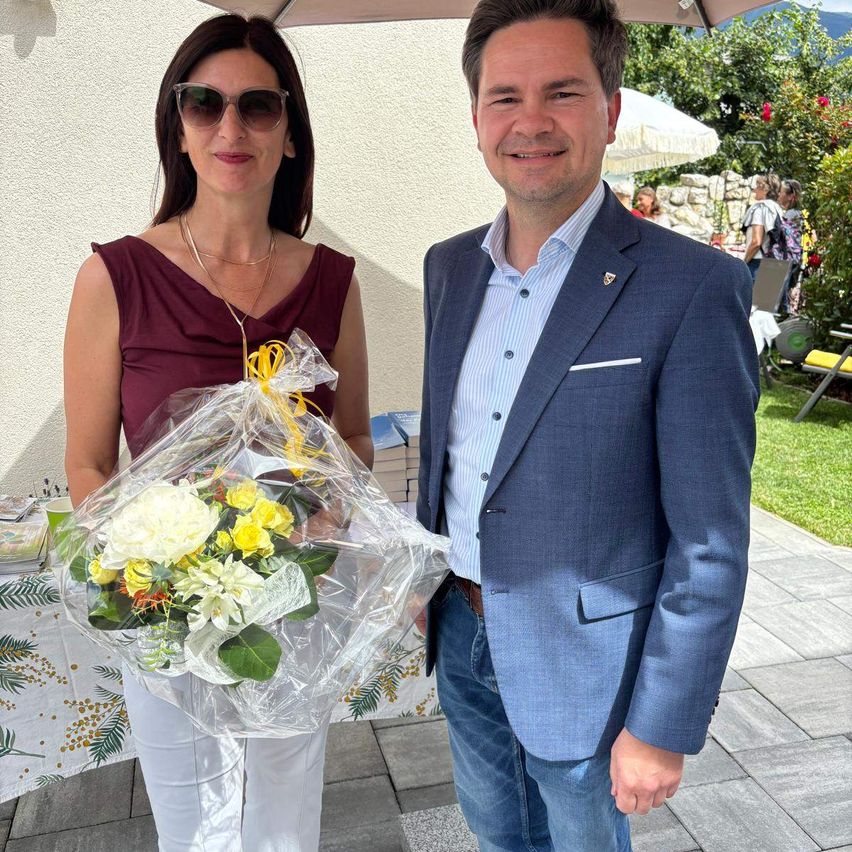 Ein Mann und eine Frau stehen unter einem Schirm. Die Frau hält einen Blumenstrauß. Sie sehen beide fröhlich aus.