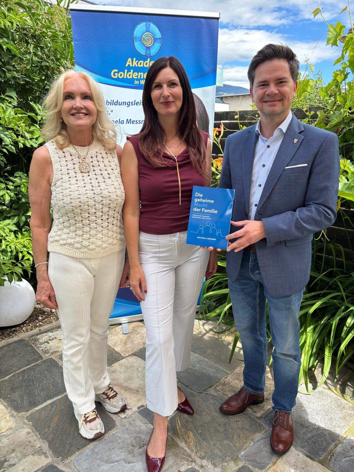 Drei Menschen stehen zusammen vor einem Garten. Ein Mann hält ein Buch mit dem Titel 'Die geheime Macht der Familie'. Die Frau links lächelt in die Kamera. Dahinter steht ein Schild mit dem Text 'Akademie Goldene in We'.