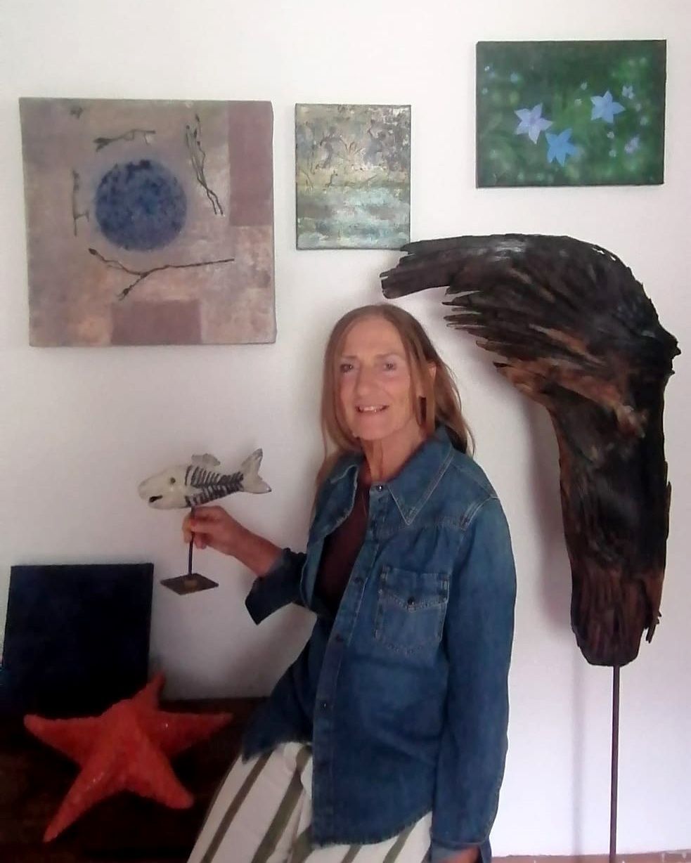 Eine Frau in einer Jeansjacke hält eine hölzerne Fischskulptur vor einer weißen Wand mit drei Gemälden.