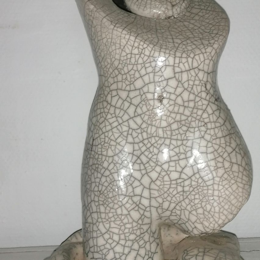Eine weiße Keramikfigur mit rissiger Oberfläche, die auf einem weißen Hintergrund sitzt und einer sitzenden Figur mit leicht gebeugter Haltung ähnelt.