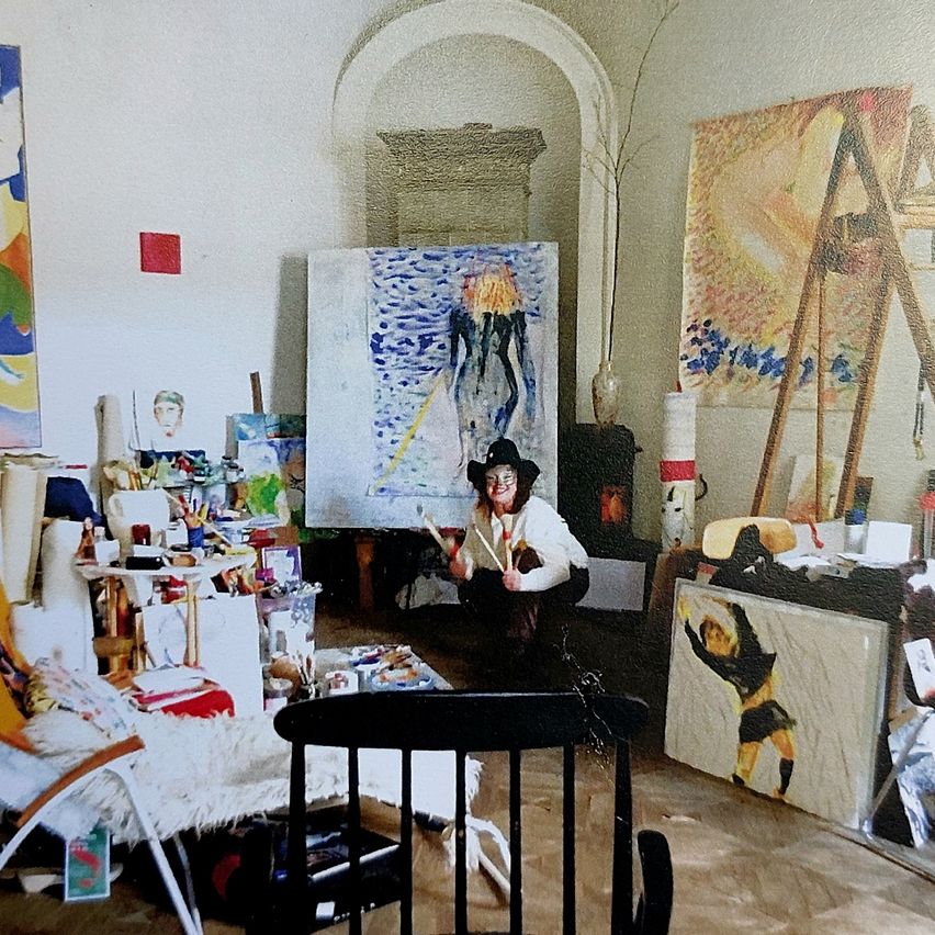 Eine Frau in einem Atelier, umgeben von Kunst. Sie trägt einen schwarzen Hut und hält Malpinsel in der Hand. Der Raum ist mit verschiedenen Gemälden, Skulpturen und Kunstmaterialien gefüllt.