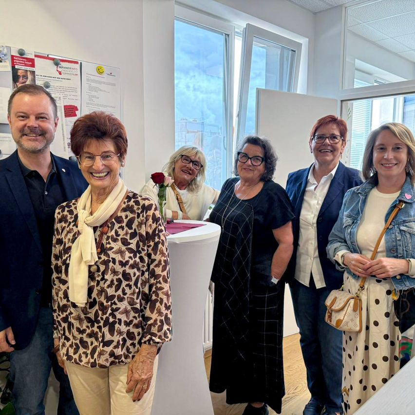 Eine Gruppe von Menschen, die zusammenstehen, lächeln und für ein Foto in einem Raum mit großen Fenstern und einem weißen Schreibtisch posieren.