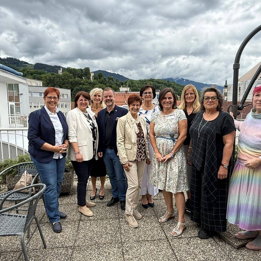 Eine Gruppe von Menschen, wahrscheinlich eine Familie, steht auf einer Terrasse und lächelt für ein Foto. Hinter ihnen sind Berge und Gebäude unter einem bewölkten Himmel.