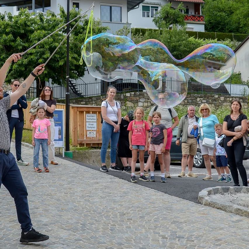 Ein Mann bläst riesige Seifenblasen für eine Menschenmenge auf einer Kopfsteinpflasterstraße, mit Häusern und Bäumen im Hintergrund.