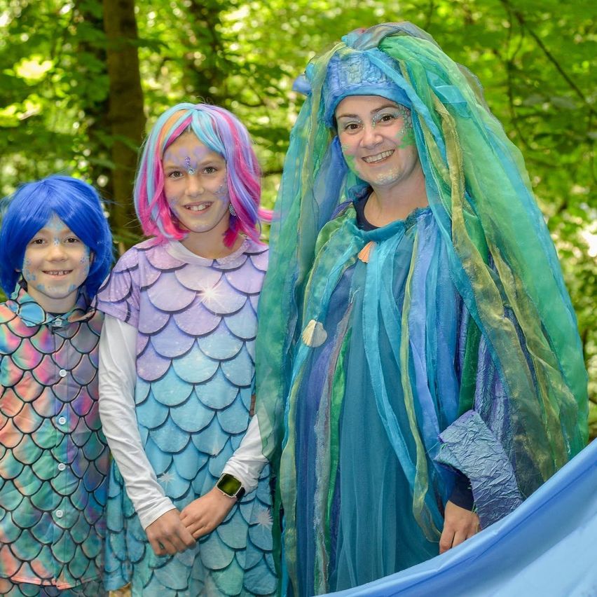 Drei Kinder und ein Erwachsener in farbenfrohen Kostümen stehen in einem Wald. Sie lächeln und posieren für ein Foto. Sie haben bemalte Gesichter und halten einen blauen Stoff.