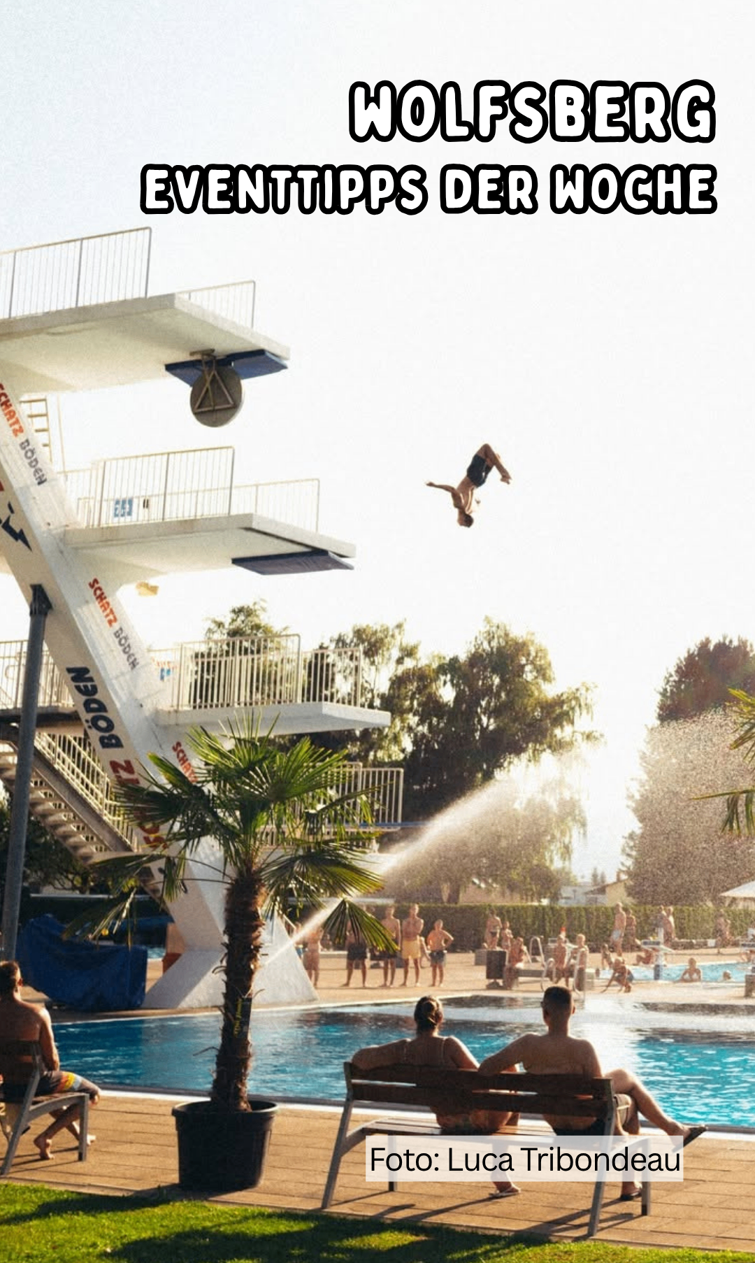 Eine Person springt von einer Sprungturm in einen Pool, wobei eine Wasserfontäne im Vordergrund sprüht, umgeben von vielen Menschen, Bäumen und einem klaren Himmel.