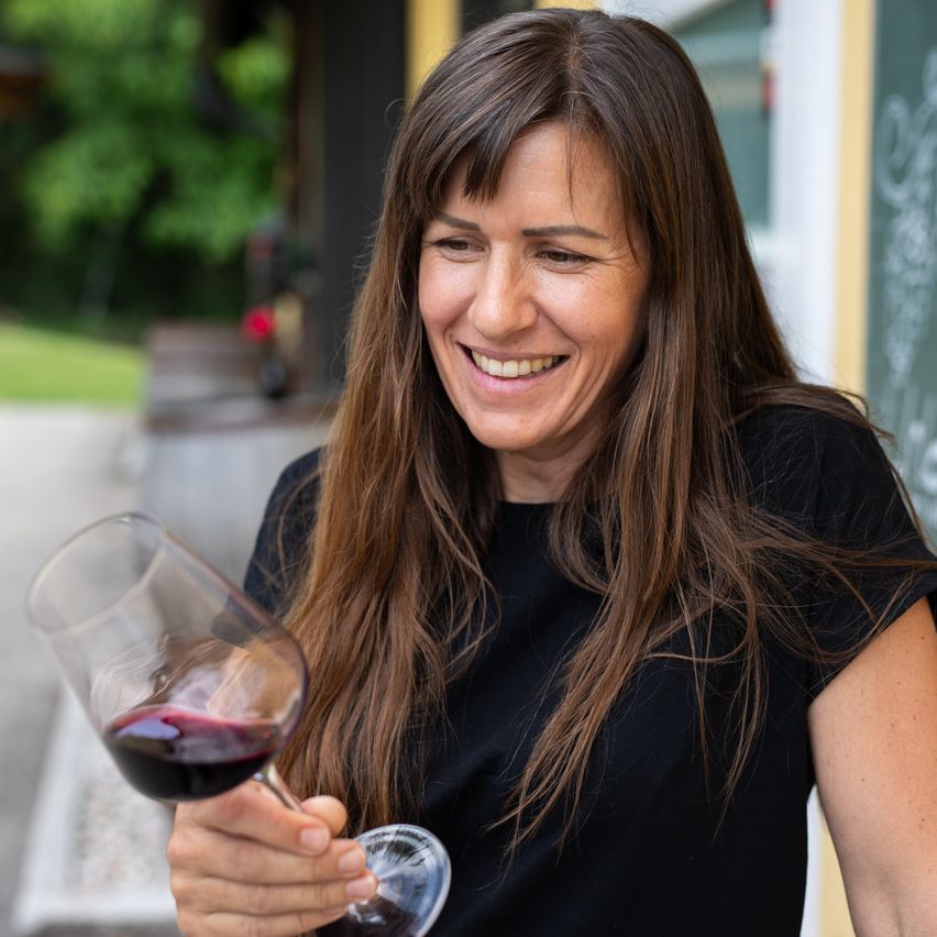 Eine Frau mit langen Haaren lächelt und hält ein Glas Rotwein in einer Außenumgebung.