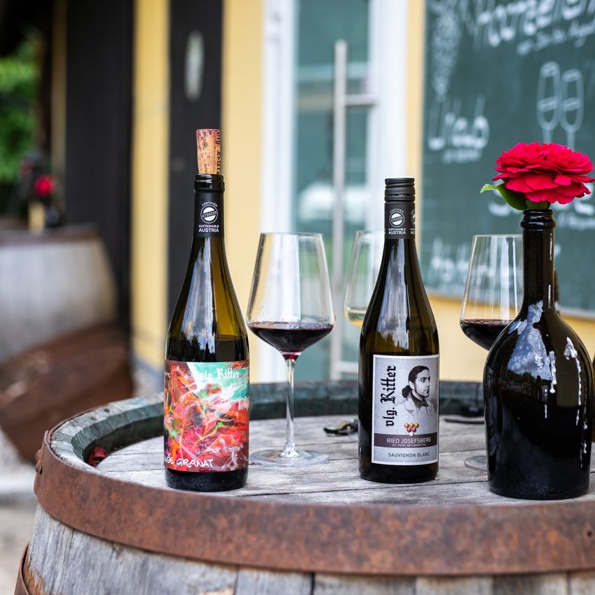 Drei Weinflaschen stehen auf einem Fass mit Weingläsern in der Nähe. Eine Flasche hat eine rote Rose darin. Die Flaschen stammen aus Österreich und heißen 'Nig Ritter'.