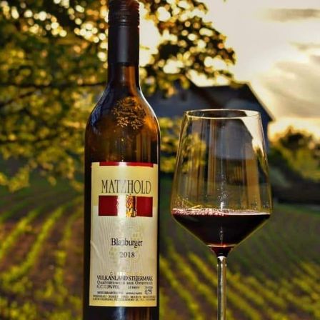 Eine Flasche Rotwein der Weinkellerei Matzhold steht neben einem Weinglas, mit Weinberg und Haus im Hintergrund.