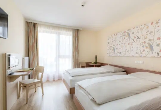 Ein Hotelzimmer mit zwei Betten, einem Schreibtisch, einem Stuhl und einem Fenster mit Vorhängen. An der Wand befindet sich ein Flachbild-TV.