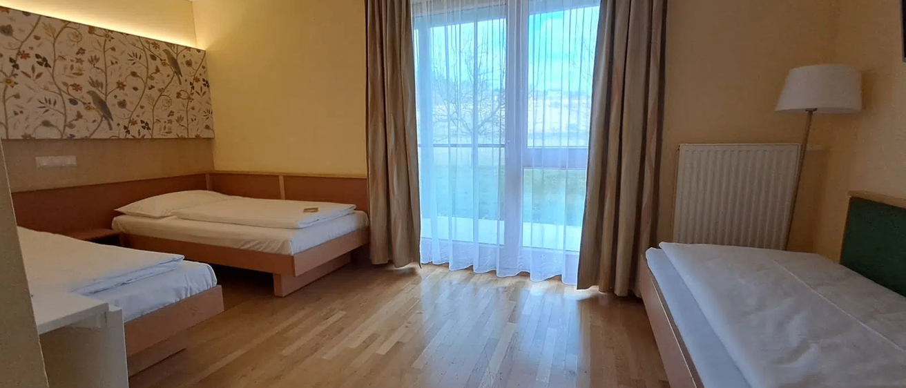 Ein Hotelzimmer mit Holzboden und einem großen Fenster mit Vorhängen. Es gibt drei Betten mit weißen Laken und Kissen.