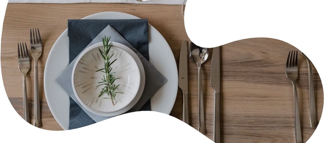 Ein elegant gedeckter Tisch mit einem weißen Teller, einem Rosmarinzweig, einem Weinglas, Kerzen und Besteck auf einer Holzoberfläche.