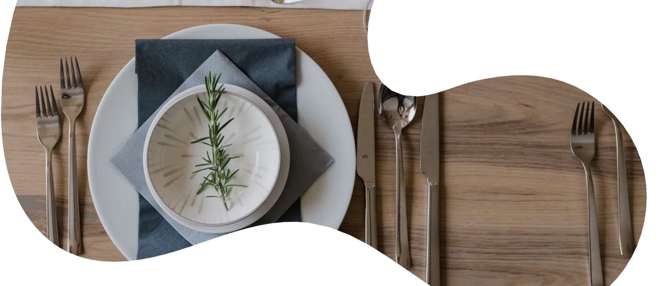 Ein elegant gedeckter Tisch mit einem weißen Teller, einem Rosmarinzweig, einem Weinglas, Kerzen und Besteck auf einer Holzoberfläche.