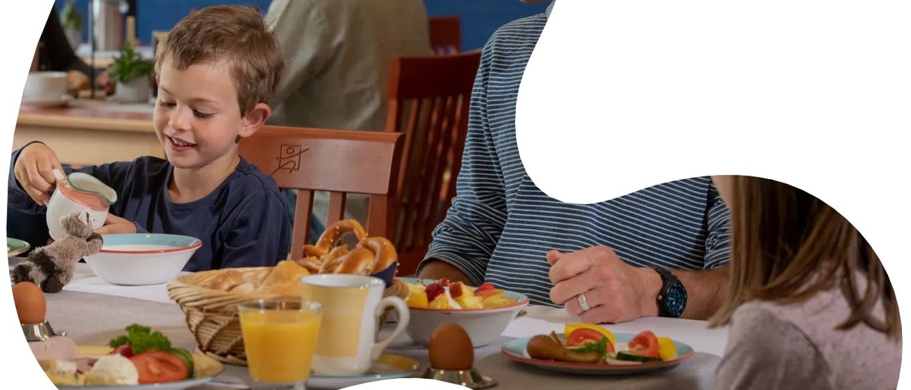 Eine vierköpfige Familie sitzt mit Essen am Tisch und lächelt. Der Mann trägt ein gestreiftes Hemd, die Frau ein graues Hemd. Der Junge hält ein Spielzeug, und das Mädchen schaut in die Kamera.