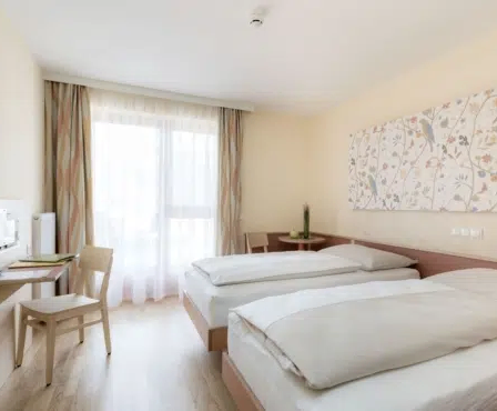 Hotelzimmer mit zwei Einzelbetten, einem Schreibtisch, einem Stuhl und einem großen Fenster mit Vorhängen. Ein Fernseher und ein Gemälde sind an der Wand angebracht.