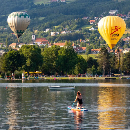 Zwei Heißluftballons schweben über einem ruhigen See mit einer Frau, die im Vordergrund auf einem Paddleboard steht. Im Hintergrund liegt eine Stadt eingebettet zwischen Hügeln, mit einer Vielzahl von Gebäuden, Bäumen und Bergen sichtbar.