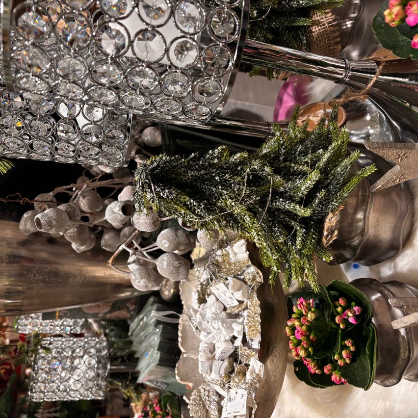 Eine dekorative Szene mit einer Kristallleuchte, Silberdekorationen, einem kleinen künstlichen Baum und rosa Blumen in einem Vasen. Die Dekorationen hängen von der Leuchte.