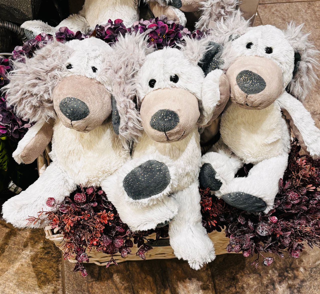 Drei weiße Plüschhunde mit grauen Ohren und Pfoten sitzen in einem Korb voller lila Blumen auf einem Holzboden.