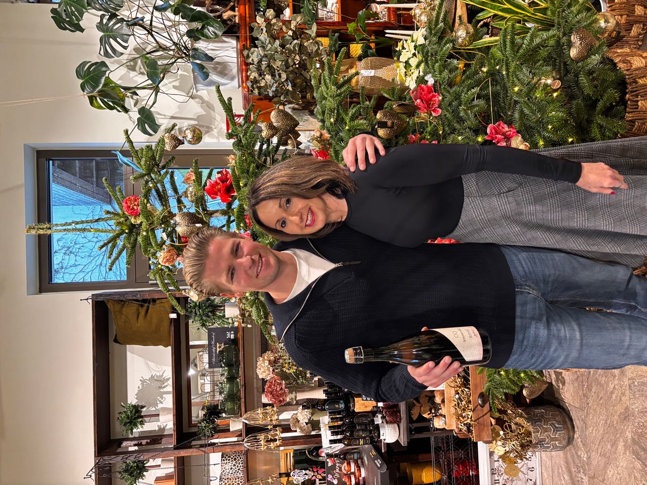 Ein Mann und eine Frau stehen lächelnd eng beieinander vor einem geschmückten Weihnachtsbaum, wobei der Mann eine Flasche Wein in der Hand hält. Dahinter befinden sich weitere festliche Dekorationen und Pflanzen.