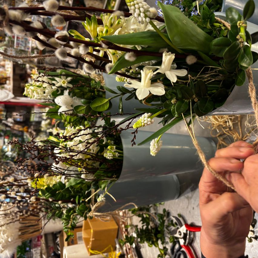 Eine Person ordnet Blumen in einer Vase an. Die Blumen sind eine Mischung aus Weiß und Grün und befinden sich in einer zylindrischen Metallvase. Die Person bindet die Blumen mit einem Seil.