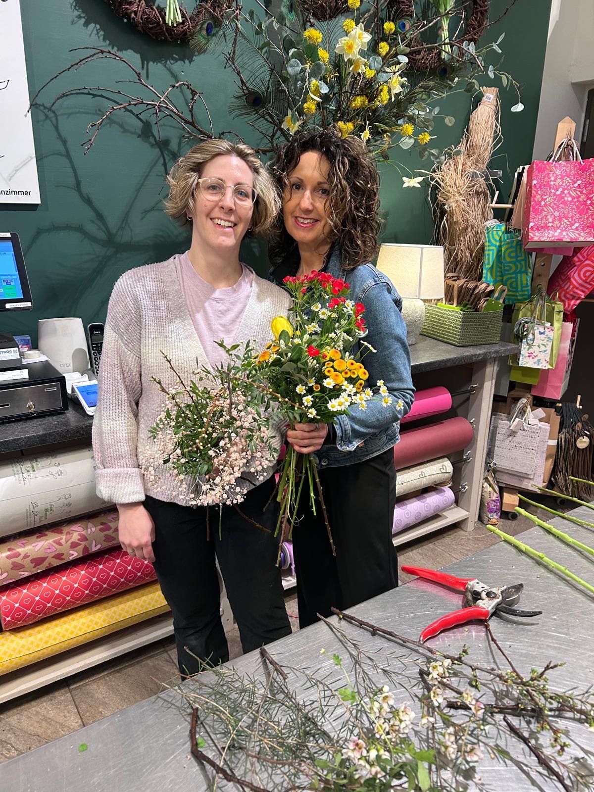 Zwei Frauen in einem Blumenladen, eine hält einen bunten Blumenstrauß und die andere hält eine Schere. Sie stehen vor einem Tisch mit Werkzeugen und eingewickelten Geschenken. Dahinter befindet sich eine grüne Wand mit einem Computermonitor und einer Lampe.