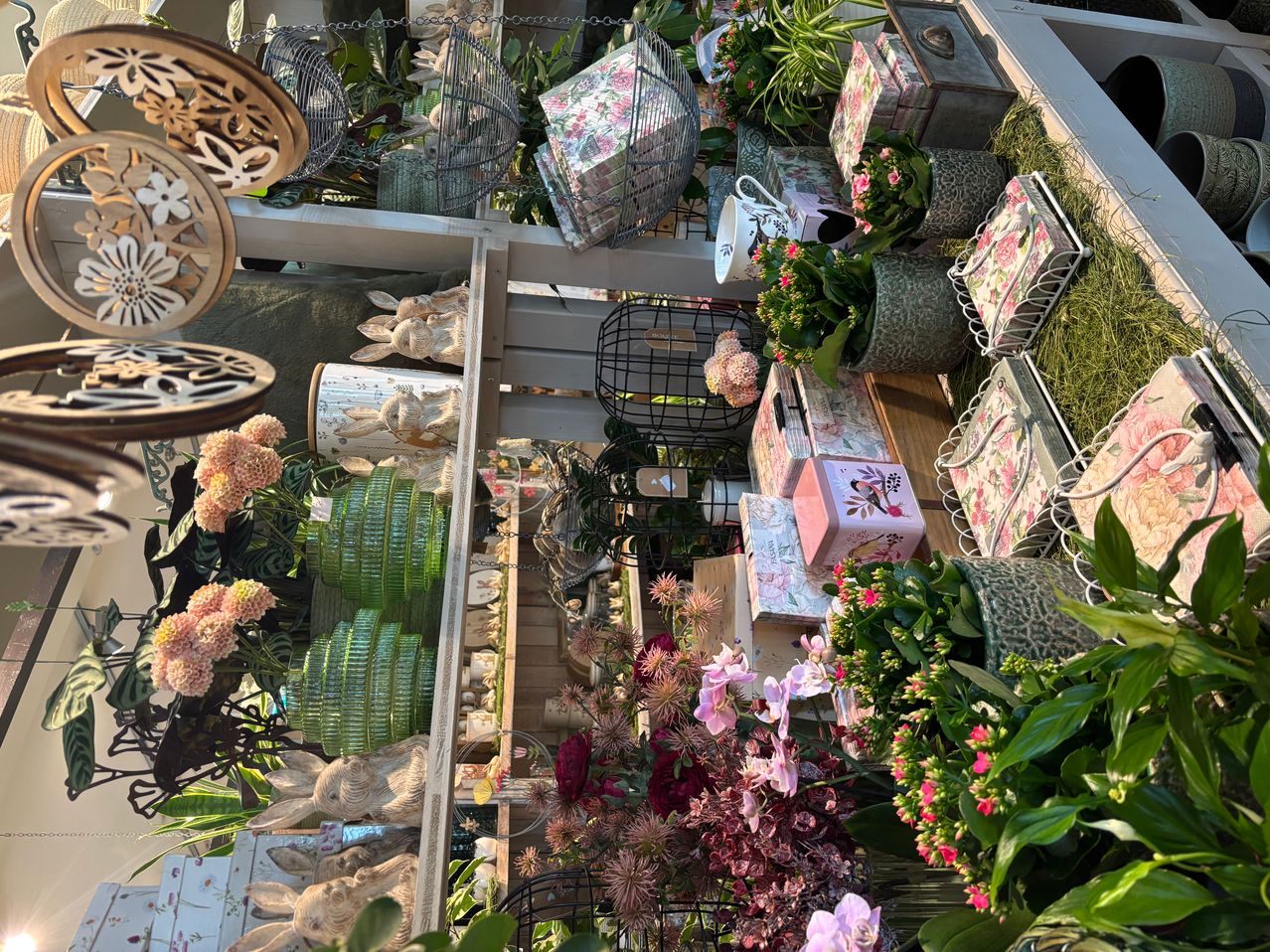 Eine Ausstellung verschiedener Blumenarrangements, Dekorationsartikel und Topfpflanzen, einschließlich rosa Blumen, grüner Vasen und Kaninchenstatuen. Die Ausstellung ist auf einer Holzplattform organisiert.
