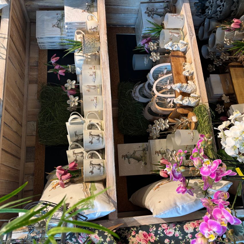 Ein Holzregal gefüllt mit verschiedenen Dekorationsartikeln, darunter Tassen, Kissen, Blumen und Pflanzen. Die Tassen haben Hasendesigns. Die Kissen haben Blumendesigns. Es gibt pinke und weiße Blumen. Das Regal hat einen Holzrahmen.