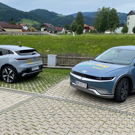 Zwei Elektroautos sind auf einem Kiesweg in einem ländlichen Gebiet geparkt, mit Bergen im Hintergrund. Ein Auto ist ein Taxi mit der Nummer 2481 an der Seite.
