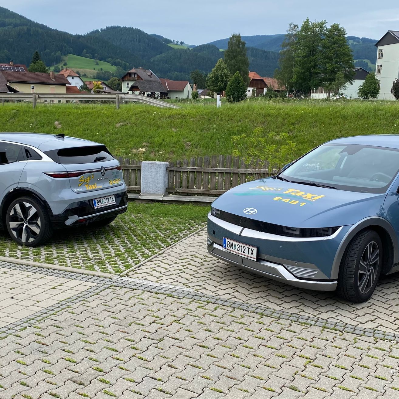 Zwei Elektroautos sind auf einem Kiesweg in einem ländlichen Gebiet geparkt, mit Bergen im Hintergrund. Ein Auto ist ein Taxi mit der Nummer 2481 an der Seite.